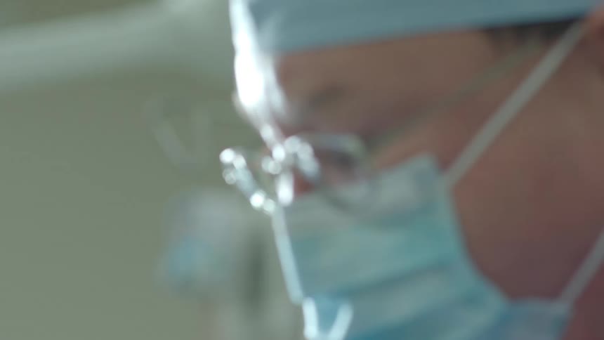A dramatic pull-focus reveals a surgeon's intense gaze during a medical procedure in the operating room