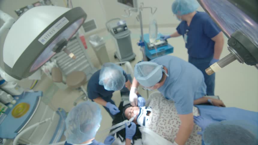 A team of surgeons in a modern operating room performs a dental procedure on a female patient, filmed from a high-angle point of view