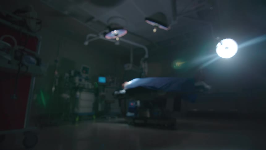 A patient lies on an operating table in a dark, empty operating room as the scene slowly goes out of focus