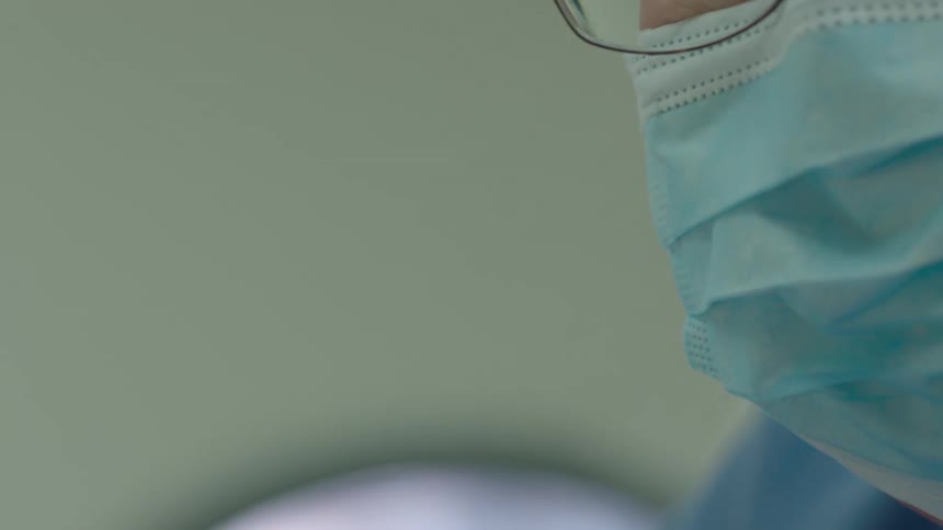 From a patient's point of view, a surgeon in a mask and glasses concentrates intently under the bright lights of the operating room