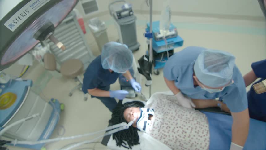 High-angle slow zoom of a medical team fitting a neck brace onto a patient on an operating table