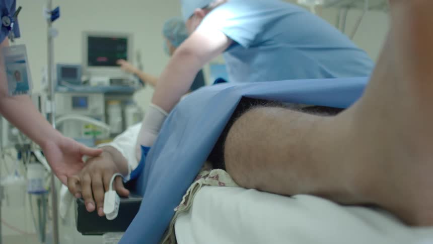 A nurse attaches a pulse oximeter to a patient's hand as the camera pulls focus to the patient's foot on the operating table