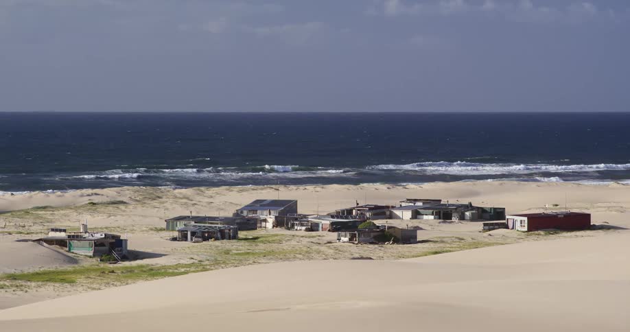 A desolate fishing village of small shacks sits on a vast sandy beach with rolling dunes as ocean waves crash ashore