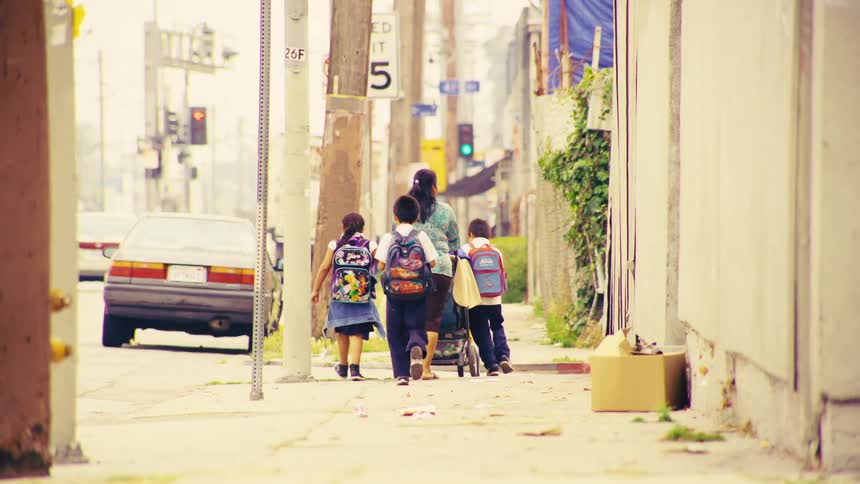 A mother and her school-aged children walk down a gritty urban sidewalk as she pushes a baby stroller