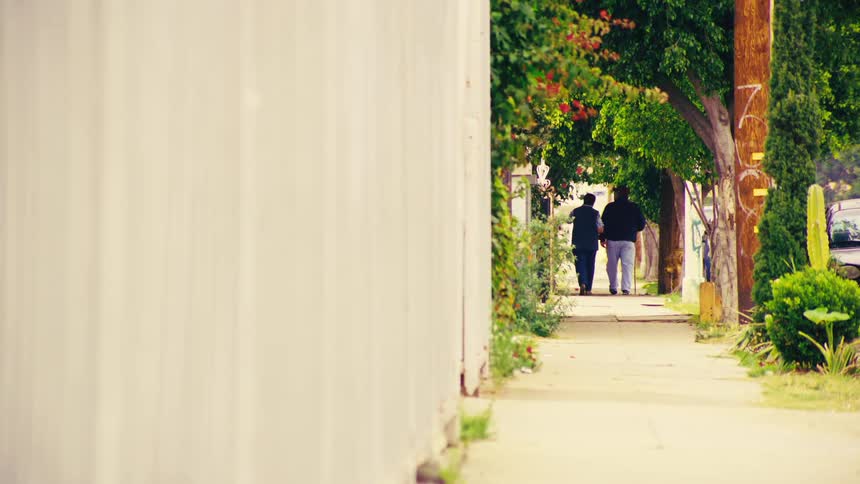 Two people take a leisurely stroll down a sidewalk lined with a white fence and lush green trees