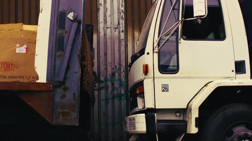 Panning shot of a gritty, industrial scrapyard with various vehicles, workers, and graffiti-covered corrugated metal walls