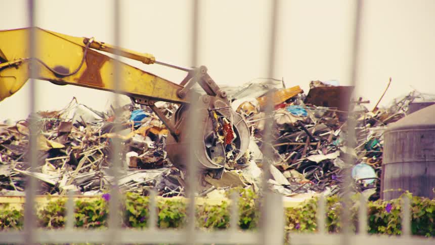 A large yellow excavator with a grabbing claw sorts through a massive pile of scrap metal at a junkyard, viewed from behind a fence