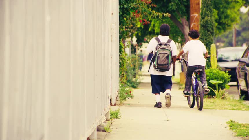 A slow-motion rear view shows two young friends, one walking with a backpack and the other on a bicycle, as they make their way down a suburban sidewalk together