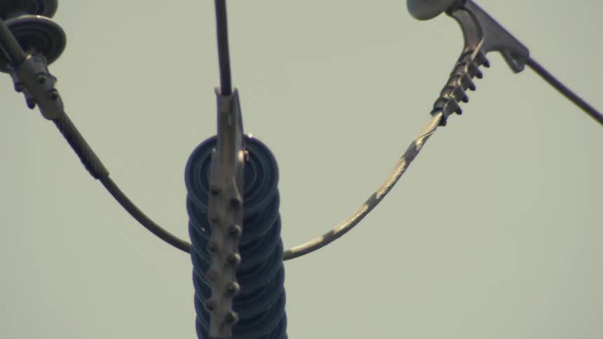 A slow, close-up pan across the intricate insulators and thick cables of a high-voltage power line against a hazy sky