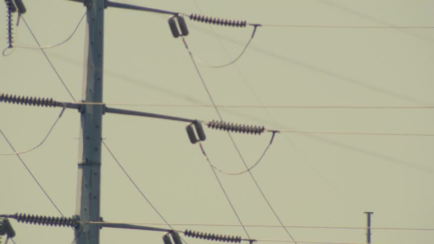 A close-up shot of high-voltage transmission towers and power lines against a pale sky, with the camera slowly tilting upwards
