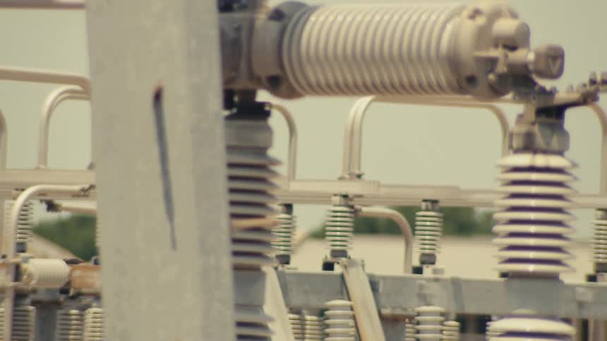 A slow-panning shot reveals the intricate details of an electrical substation with a city skyline blurred in the background
