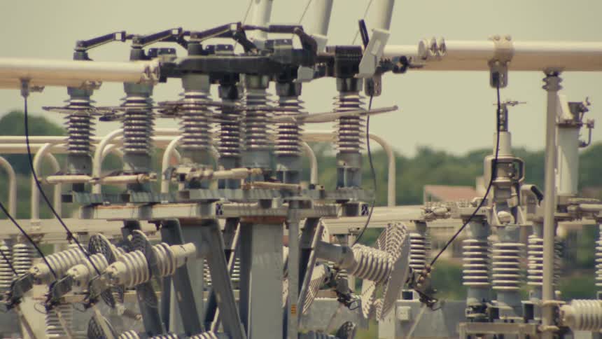 A close-up slow pan across the intricate network of transformers and insulators at a power substation on a hot day