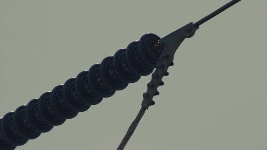 A detailed close-up shot pans along high-voltage power lines and insulators against a grey sky