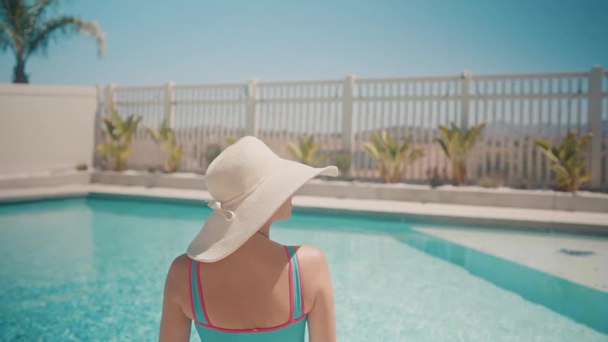 A woman wearing a wide-brimmed sun hat relaxes by a crystal clear swimming pool on a sunny summer day
