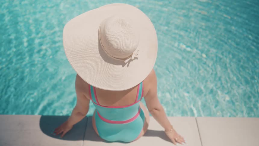 A woman in a stylish sun hat and swimsuit relaxes by the edge of a serene swimming pool on a sunny day