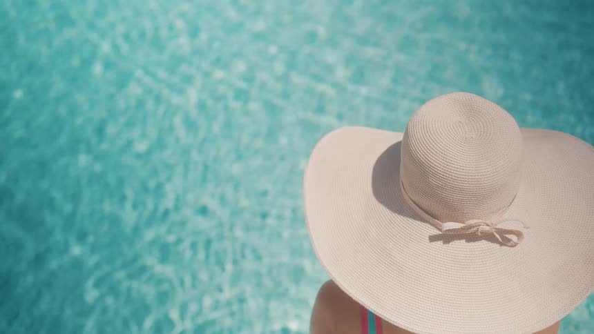An overhead view of a woman in a large white sun hat sitting by the tranquil, sparkling turquoise water of a swimming pool