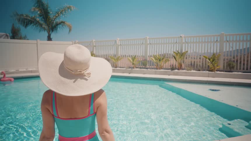 A stylish woman in a large sun hat and colorful swimsuit enjoys a tranquil, sunny day by her backyard pool