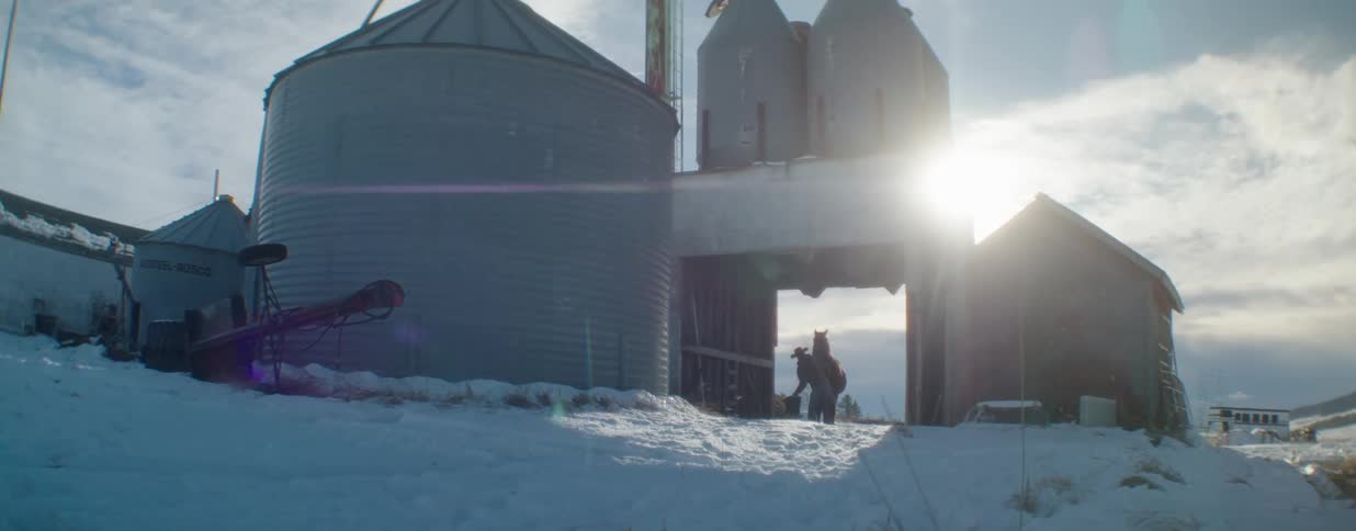 A rancher leads their horse and dog through a snowy farmyard past large grain silos at sunrise