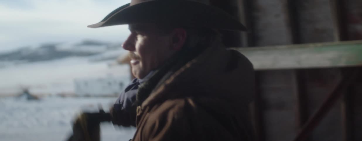 A cowboy wearing a brown jacket and hat mounts his horse inside a barn and looks out at the snowy landscape