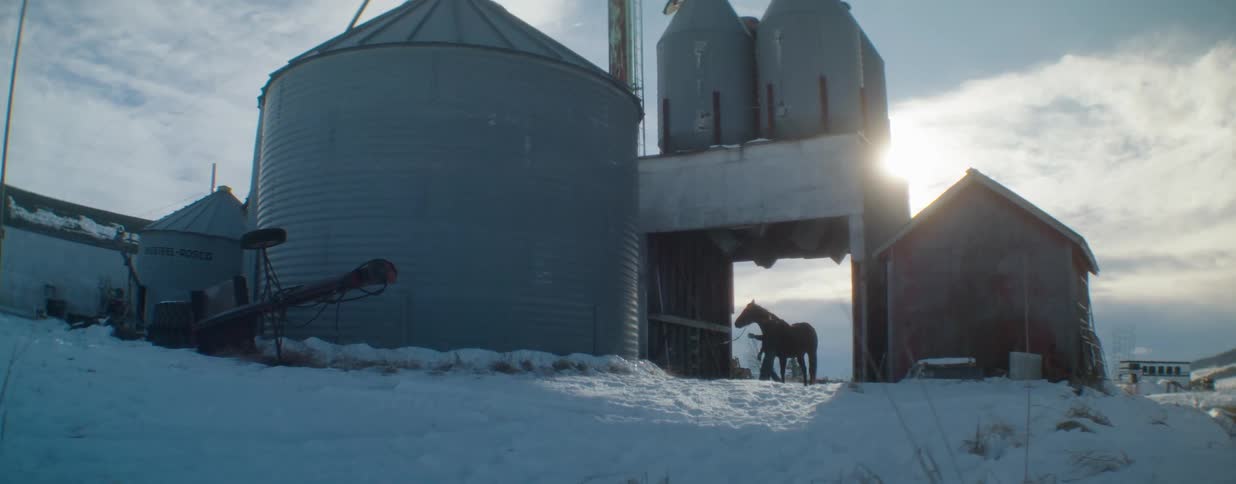 A silhouetted rancher and his horse stand between large grain silos on a farm covered in snow during a cold winter day