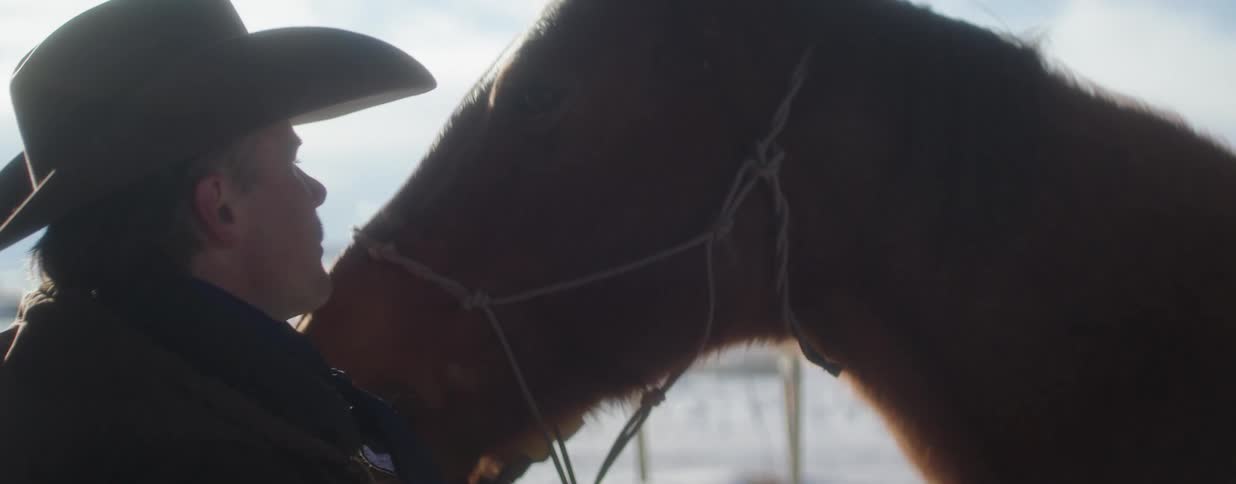 An intimate close-up of a cowboy in a hat affectionately petting his brown horse in a snowy winter landscape