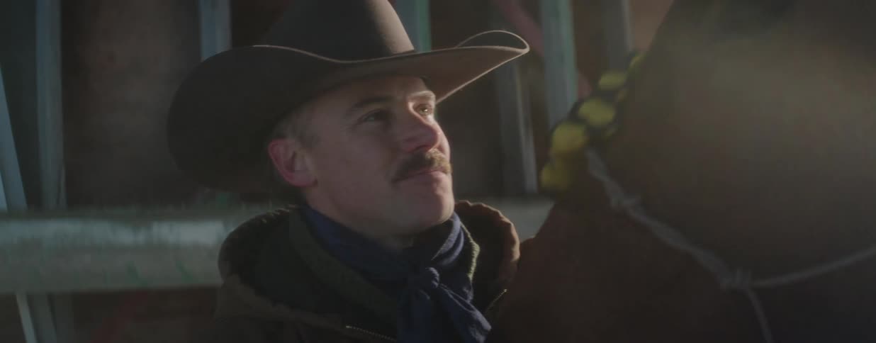 A thoughtful cowboy stands by his horse in a rustic barn, illuminated by a soft, warm light