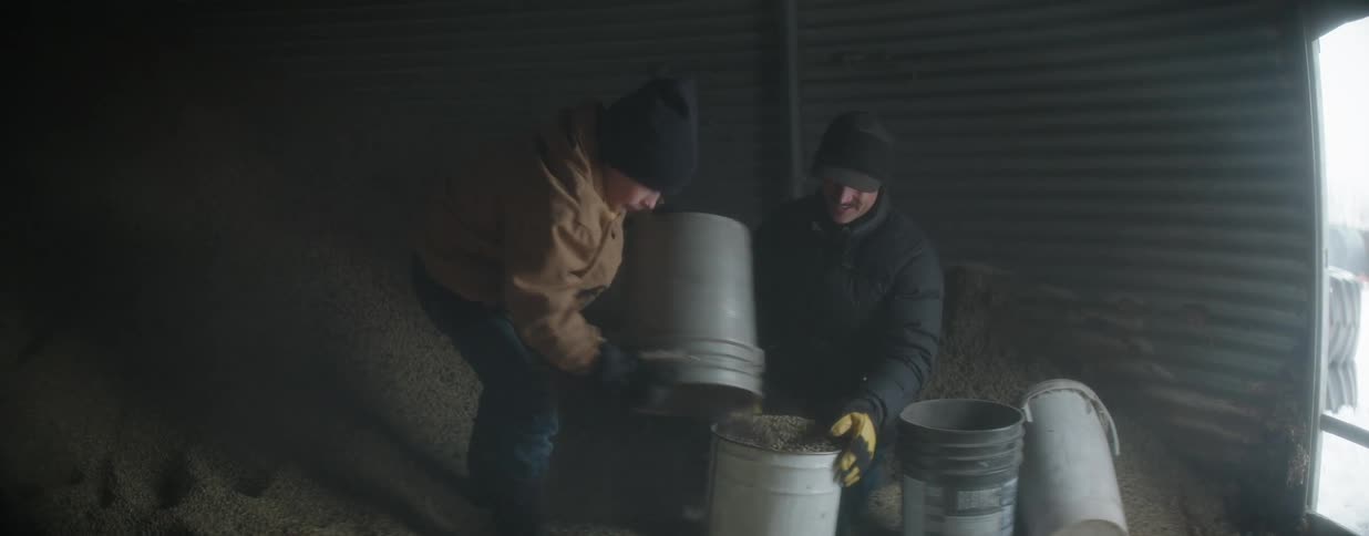 A young boy helps his father fill buckets with grain inside a large, dusty grain bin