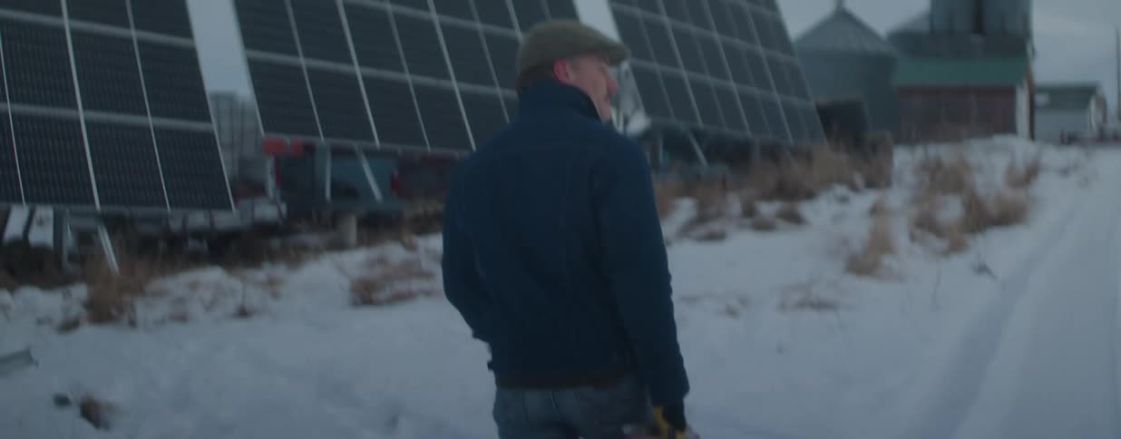 A man carries a gas can as he walks through the snow toward a large solar panel array on a rural farm