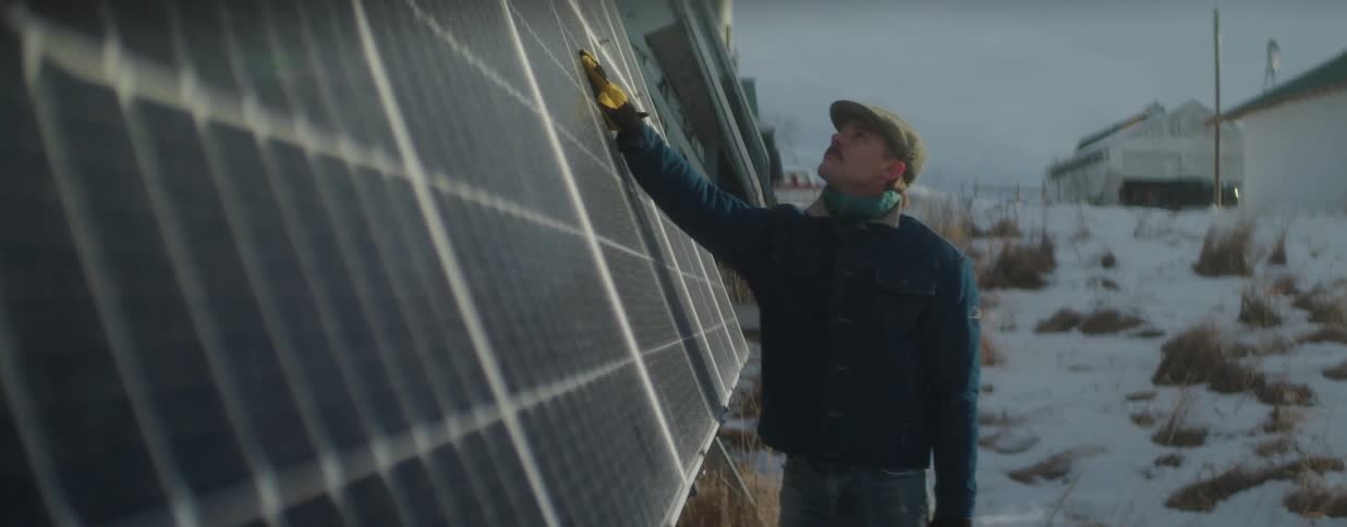 A man wearing a hat and gloves inspects solar panels in a snowy field on a farm