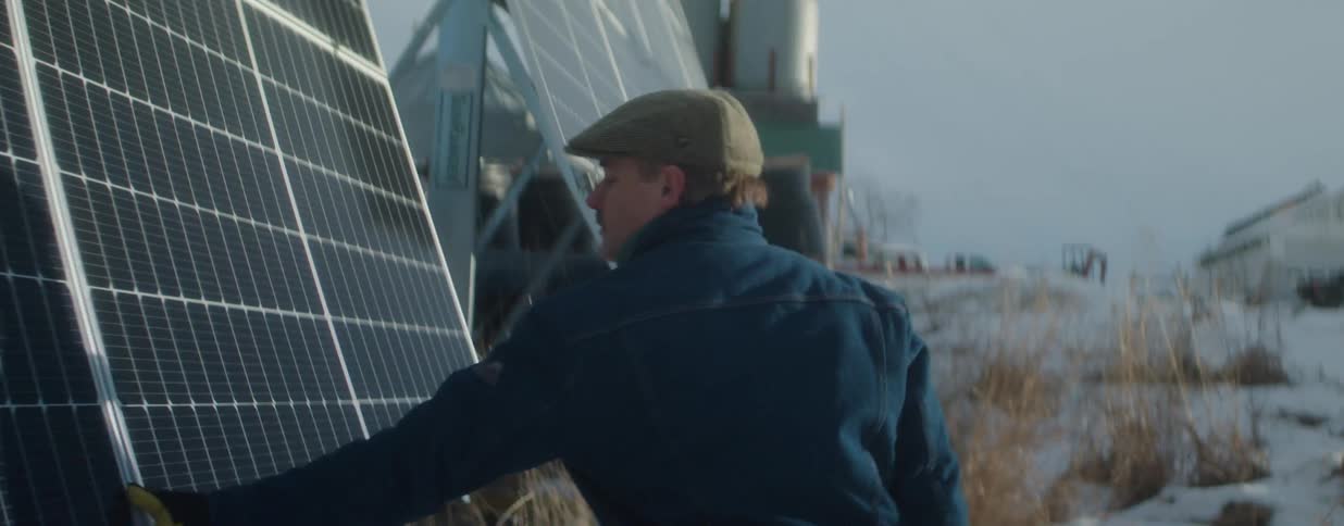 A man in a cap and denim jacket inspects his solar panel array on a snowy rural farm