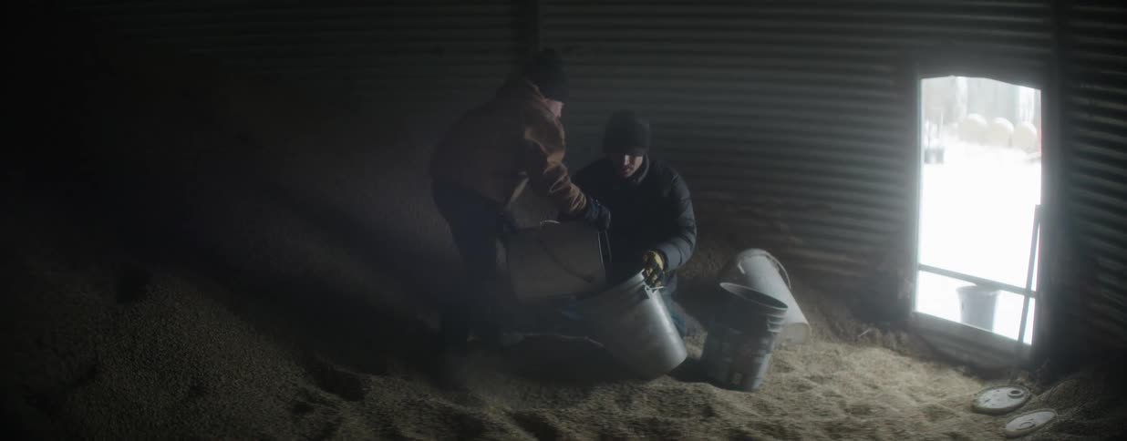 Two farmers work together in slow motion scooping grain into buckets inside a dimly lit and dusty silo