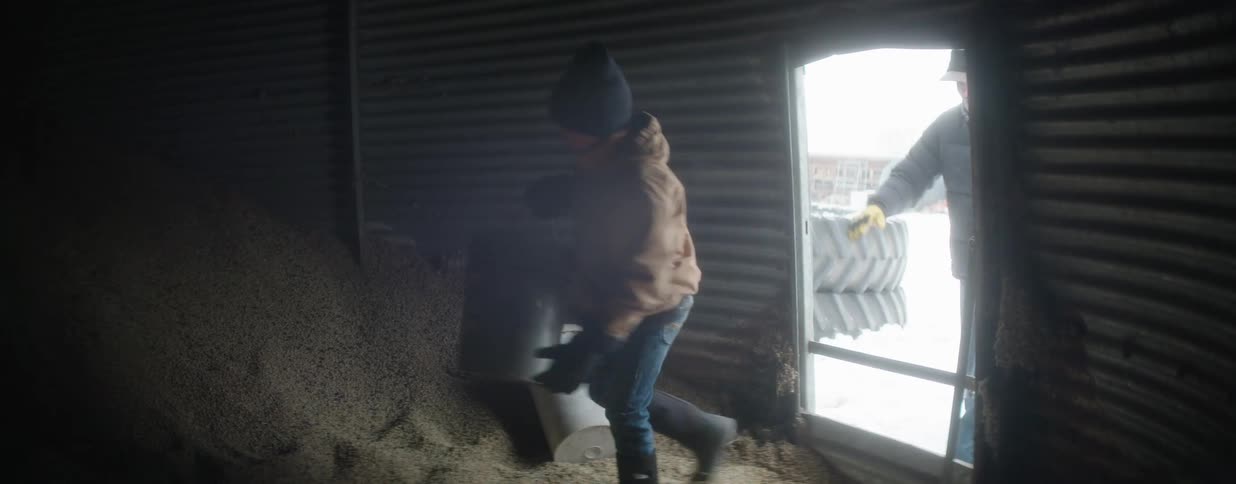 A father and his son work together in slow motion, entering a dark grain silo to fill up buckets with animal feed on a cold winter day