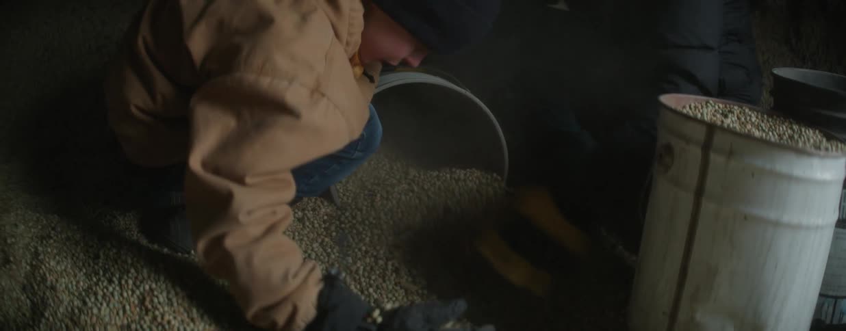A dedicated father and his son work together inside a dark grain bin, scooping feed by hand into a bucket