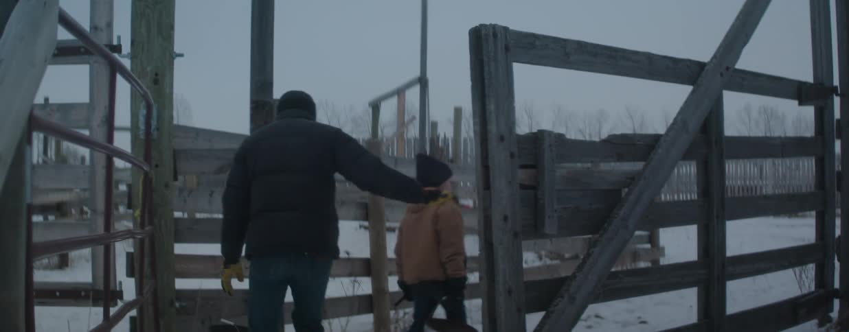 A father and son walk through a wooden gate with buckets to do chores on a snowy farm in slow motion