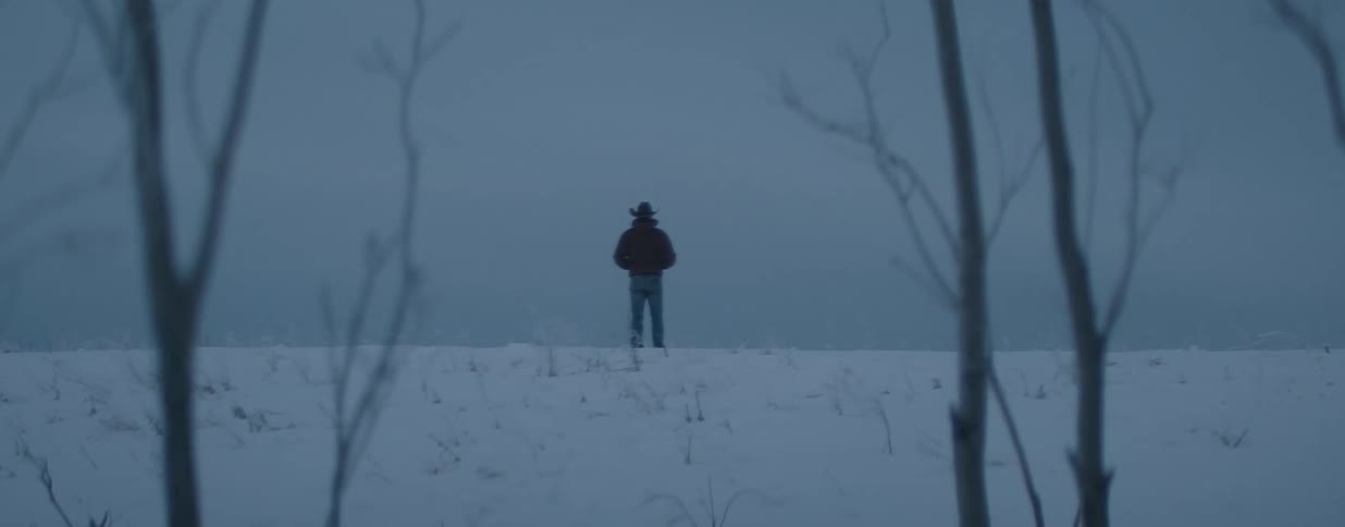 A cowboy stands with his back to the camera, looking out over a desolate, snow-covered field at dusk