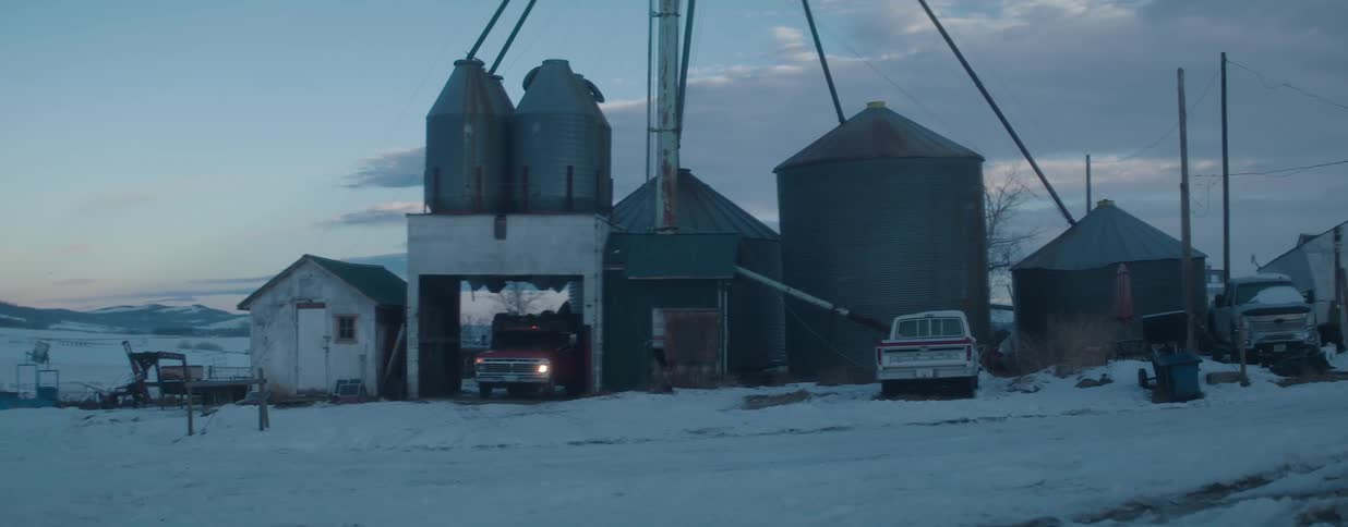A worker walks across a snowy farmyard, gets into a large red truck, and drives it away from the silos at dawn