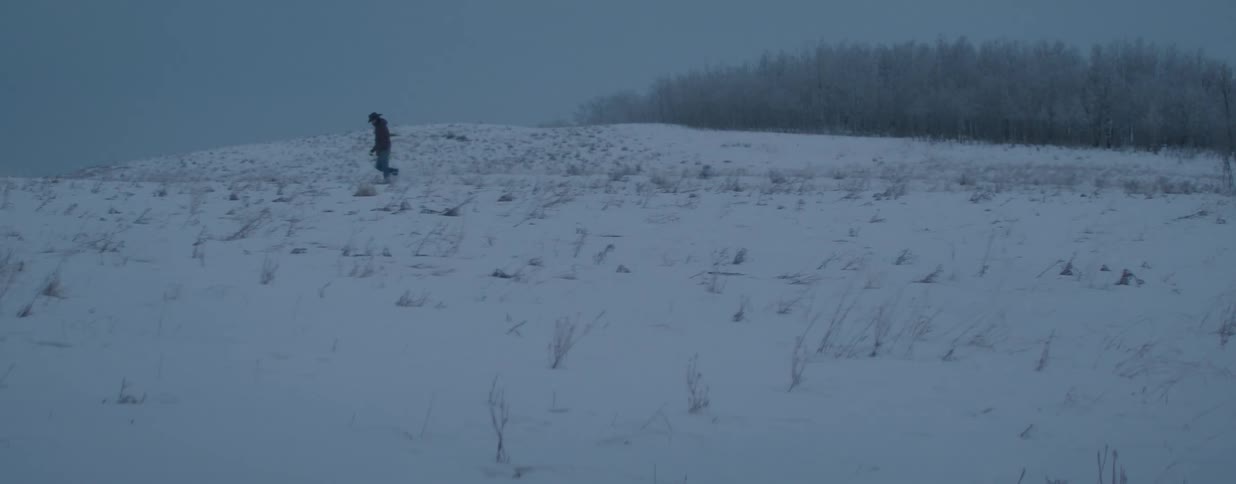 A lone cowboy walks across a vast, snow-covered landscape during a bleak winter dusk