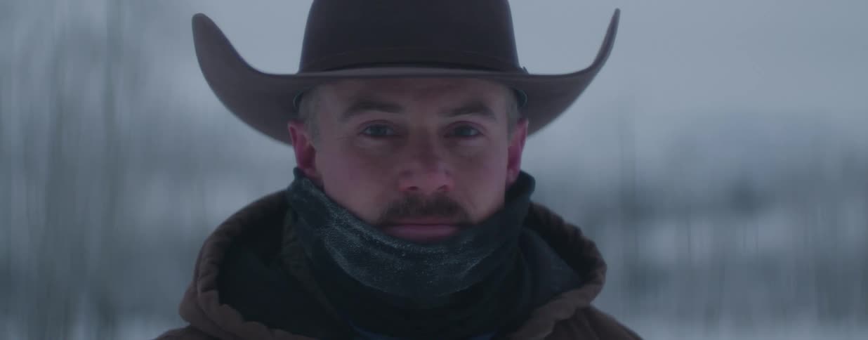 A stoic cowboy bundled up against the cold looks intently at the camera as snow falls around him