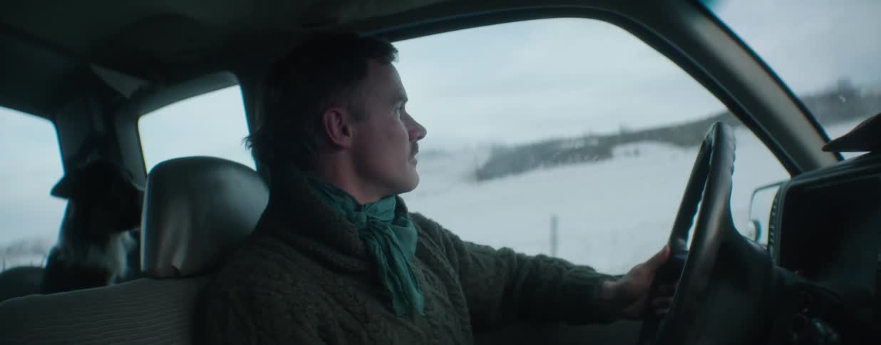 A man with a mustache and a green neckerchief drives thoughtfully through a snowy rural landscape