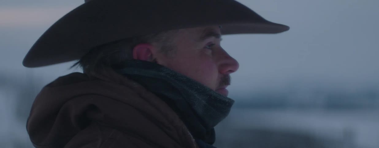 A stoic cowboy wearing a hat and warm winter coat gazes off into the distance on a cold, overcast day