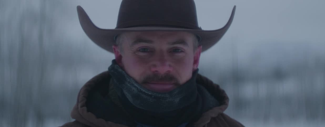 Close-up portrait of a mustached cowboy in a hat and frosted scarf, smiling gently in a snowy winter setting