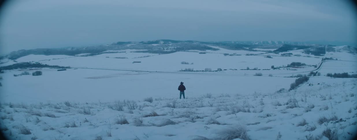 A lone cowboy walks across a snowy hill and stops to survey the vast, frozen landscape below at dusk
