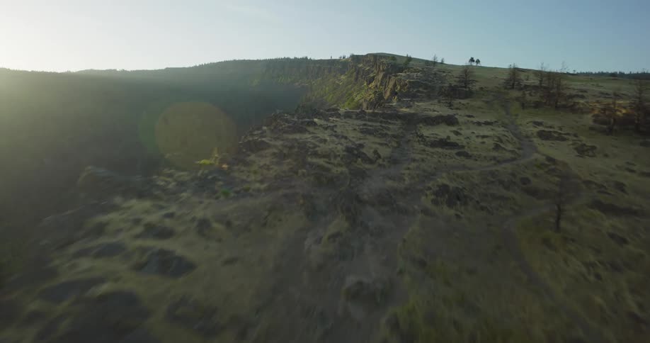A breathtaking aerial shot soars along the edge of a massive rocky bluff overlooking a forested valley at sunrise