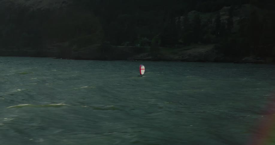 Cinematic aerial shot follows a lone wing foiler gliding across a vast, choppy river at dusk with mountains on the horizon