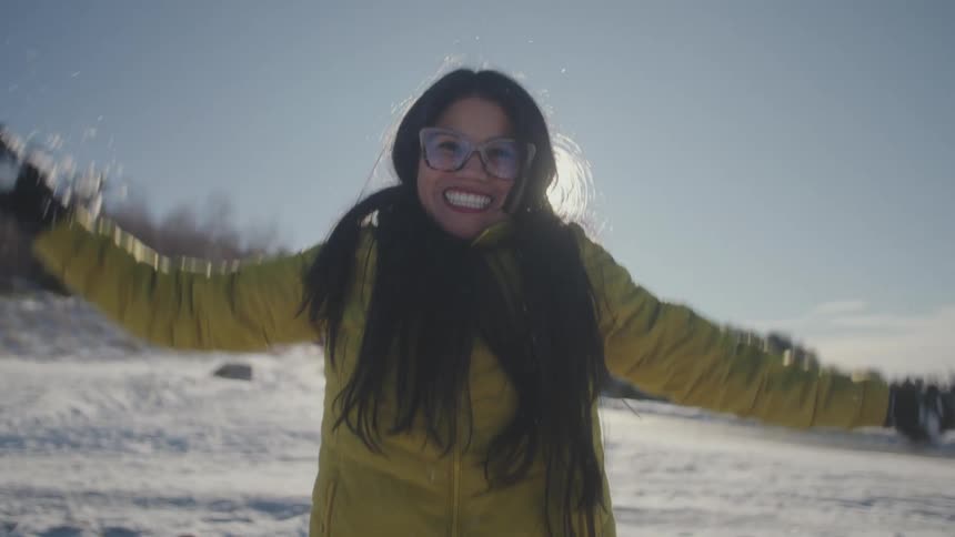 A smiling woman wearing a yellow jacket joyfully throws snow up into the air in slow motion on a bright winter day