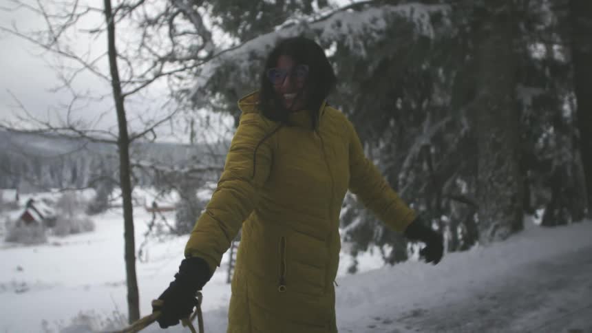 A smiling woman in a yellow winter coat holds onto a sled rope while standing in a snowy forest in slow motion