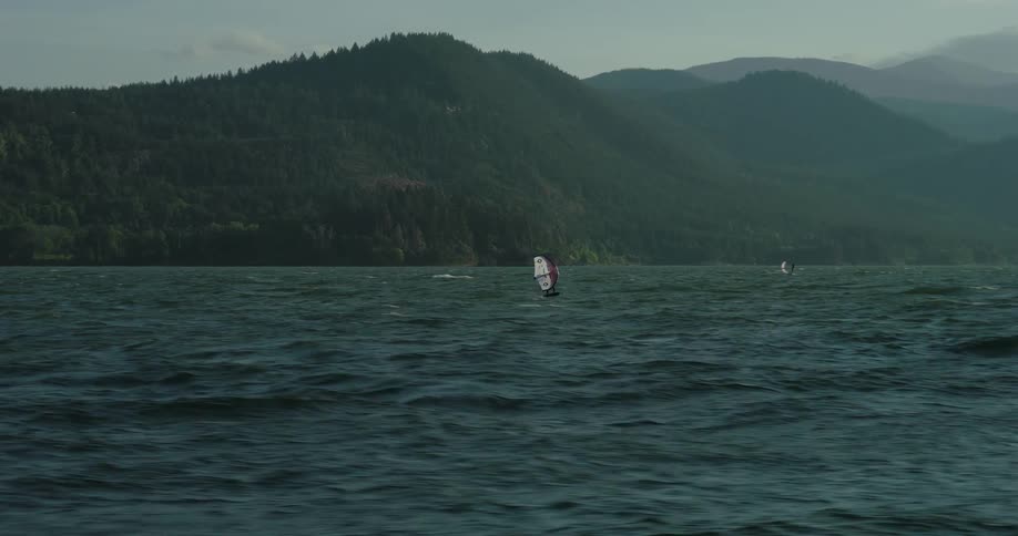A wing foiler gracefully glides across the choppy water of a river gorge in slow motion, with majestic tree-covered mountains in the background