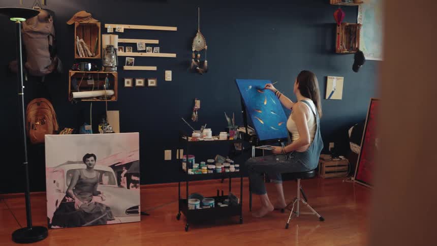 A reveal shot shows a young woman painting a vibrant school of fish on a canvas in her artistic home studio