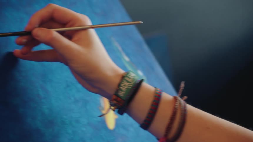 A close-up shot of a young female artist's hand as she carefully paints a whale onto a blue canvas in her studio