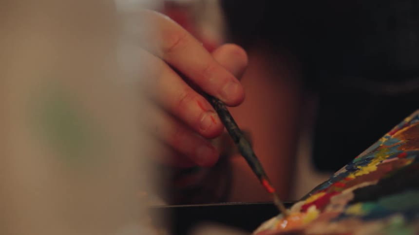 A young female artist in paint-splattered overalls focuses intently as she mixes colors on her palette in a warmly lit studio